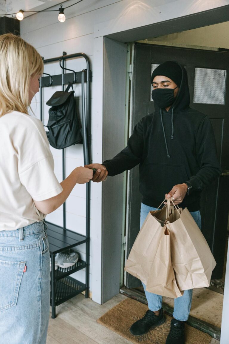 A masked courier hands over shopping bags to a customer in a contactless delivery exchange.