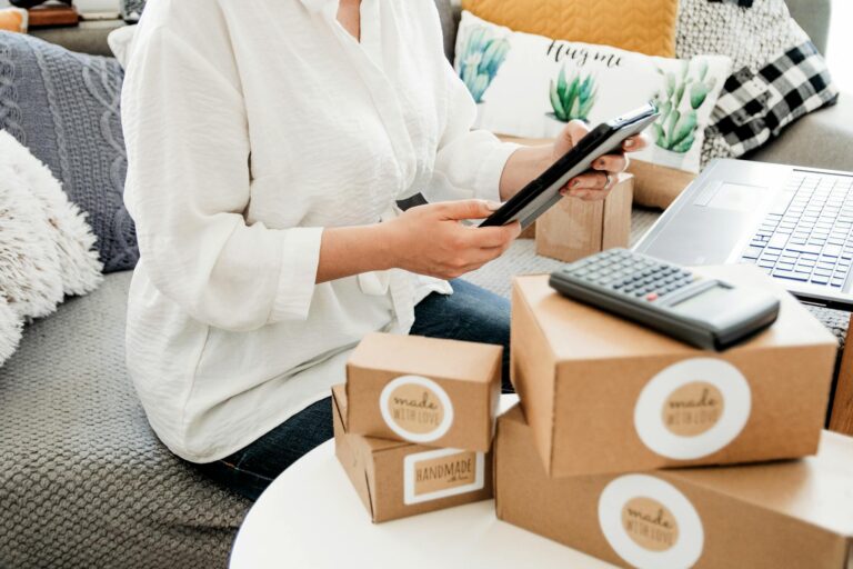 Woman in white blouse handling online business orders with tablet, boxes, and calculator.